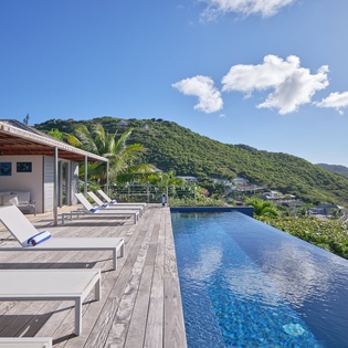 Stunning infinity pool overlooking lush tropical mountains and sparkling ocean waters, with premium sun loungers on expansive wooden deck.