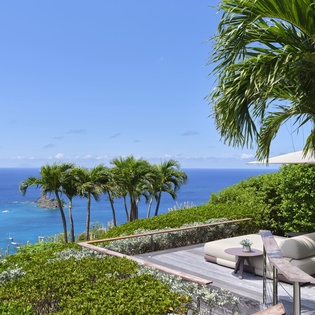 The property features a terraced outdoor area with stunning ocean views, surrounded by tropical palms and lush greenery, offering a serene setting overlooking crystal-blue waters.