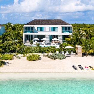Modern beachfront villa with pristine white sand beach and crystal-clear turquoise waters, surrounded by lush tropical palms and vegetation.