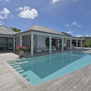 A contemporary villa showcases its sleek covered terrace and pristine swimming pool, surrounded by tropical landscaping under bright skies.