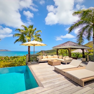 Stunning infinity pool overlooking turquoise Caribbean waters, with plush loungers, shaded seating under swaying palms, and panoramic coastal views. Your private paradise awaits.