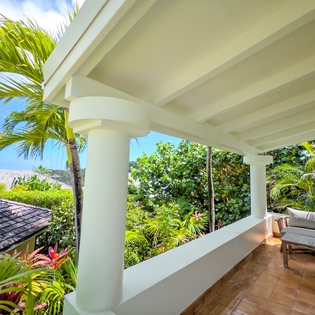 A covered balcony framed by white columns and lush tropical palms offers a serene spot to relax with ocean glimpses through the greenery.