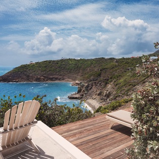 A secluded coastal hillside offers a dramatic panoramic view of turquoise waters, rugged cliffs, and lush green vegetation stretching toward distant villas under a Caribbean sky.