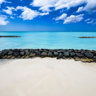 Pristine white sand beach with turquoise waters and natural rock formations, framed by dramatic blue skies and fluffy clouds in the surrounding area.