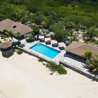 Aerial view showcasing a beachfront vacation property with private pool, multiple structures, and lush tropical landscaping set along pristine white sand and dense foliage.