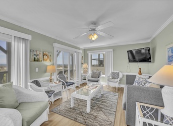 Your bright, coastal-style living room welcomes you with plush seating, soothing colors, and natural light streaming through doors opening to scenic views beyond.