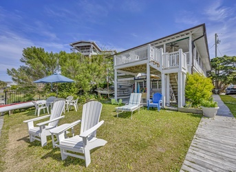 Charming two-story vacation home featuring a spacious lawn with Adirondack chairs and covered porch area surrounded by mature trees.