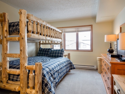 Your cozy mountain bedroom features handcrafted log bunk beds with plaid linens, a dresser with TV, and warm lighting—perfect for families seeking alpine charm and comfort.
