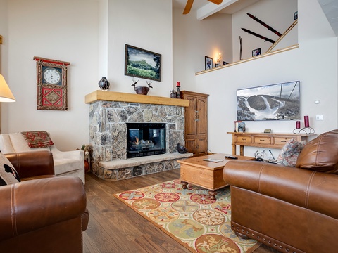 Unwind in your spacious living room with vaulted ceilings, a cozy stone fireplace, and plush leather seating. Natural light streams through the windows as you relax near the warmth of the fire, surrounded by mountain-themed décor that brings the outdoors i