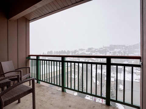 A covered balcony overlooks a snowy neighborhood and distant hills, offering a peaceful winter retreat with cozy seating for two.