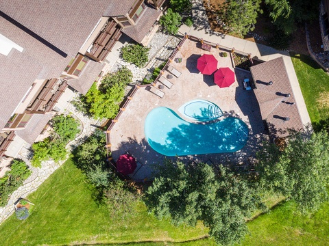 Aerial view showcases a stunning kidney-shaped pool surrounded by lush lawns, vibrant umbrellas, and comfortable sun loungers, all nestled within beautifully landscaped gardens beside the main building.