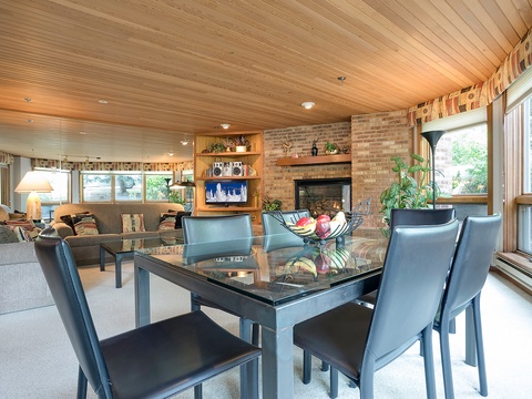 Unwind in this warm, open-concept space where natural wood ceilings and brick fireplace create a cozy retreat. Enjoy meals at your dining table while soaking in the tranquil greenery through expansive windows.