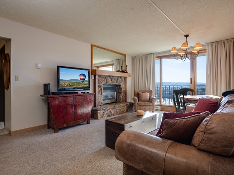 Settle into your cozy living room where plush seating faces a warm stone fireplace, and sunlight streams through doors opening to your private balcony with stunning views beyond.