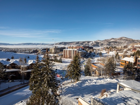 A snow-covered resort town nestled between mountains and lake offers scenic winter views under a clear blue sky.