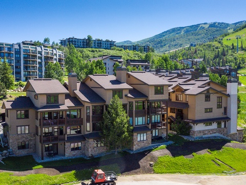 Multi-level mountain resort buildings nestled in a scenic valley setting, surrounded by lush greenery and rolling hills with ski slopes visible in the distance.