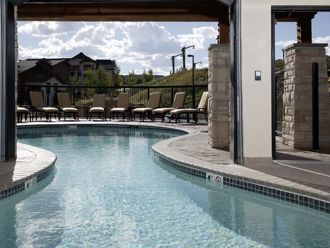 A winding outdoor pool surrounded by lounge chairs offers a peaceful setting with mountain views and neighboring homes, accessed through a shaded stone-framed entrance.