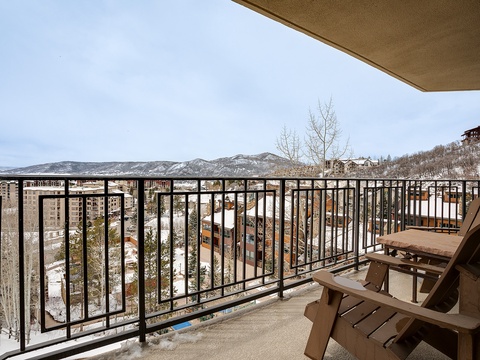 A mountain resort balcony overlooks snow-dusted slopes and neighboring buildings, framed by distant peaks under a soft winter sky.