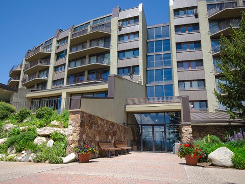 Modern multi-story property nestled into a landscaped hillside, featuring multiple balconies and a welcoming stone-accented entrance surrounded by vibrant gardens and greenery.