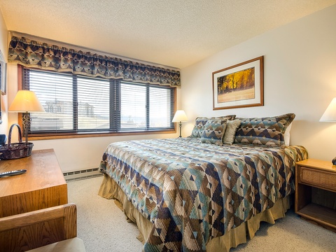 Your cozy bedroom features a plush queen bed with rustic quilted bedding, warm bedside lamps, and large windows that fill the room with natural light and peaceful views.