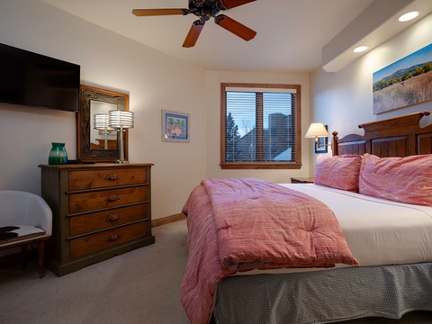 Your cozy bedroom features a plush bed with soft linens, warm wood furnishings, a ceiling fan, flat-screen TV, and natural light from a window with mountain views.
