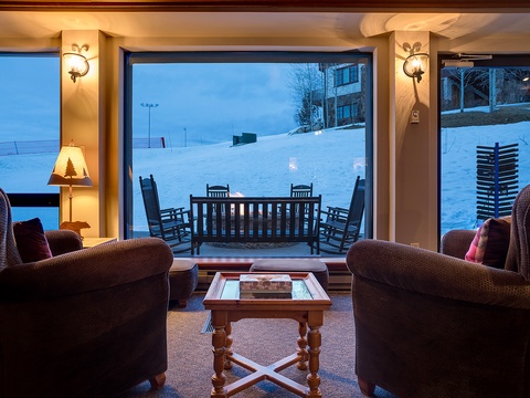 Curl up in plush seating as you gaze through expansive windows framing a serene winter wonderland, where gentle lamplight creates a warm, inviting glow against the snowy landscape outside.