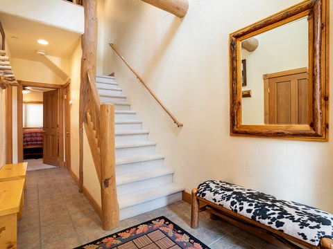 Step into your cozy alpine retreat where rustic log beams and warm wood tones create an inviting mountain atmosphere. The bright staircase leads you to discover more comfortable living spaces above.