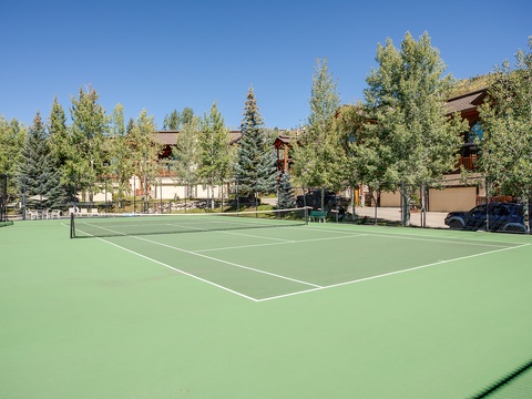 A pristine tennis court surrounded by mature evergreens and mountain-style buildings, offering guests an active outdoor amenity in a tranquil alpine setting.