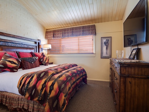 Unwind in your cozy retreat featuring a plush bed dressed in vibrant Southwestern-style linens, rustic wood furnishings, and a warm vaulted ceiling that invites relaxation.
