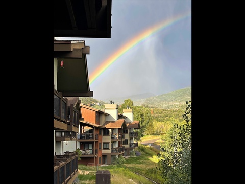 A vivid double rainbow arcs over the property's wooded mountain setting, with multi-level residential buildings nestled in the forested valley below.