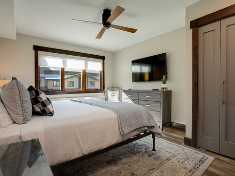 Your bright and restful bedroom awaits, with natural light pouring through large windows, a plush bed dressed in soft linens, and a TV for your evening unwind. Modern comfort meets warm wood tones.