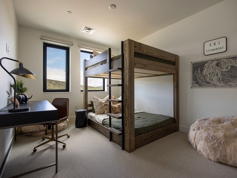 This cozy bedroom features custom bunk beds with rustic wood framing, perfect for families or groups. Natural light streams through large windows, highlighting the minimalist workspace and soft textures throughout.