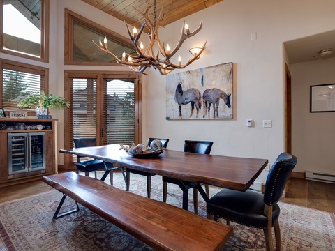 Gather around this handsome dining table beneath a dramatic antler chandelier. Natural light floods in through tall windows, highlighting the rustic elegance and inviting warmth perfect for your mountain getaway.