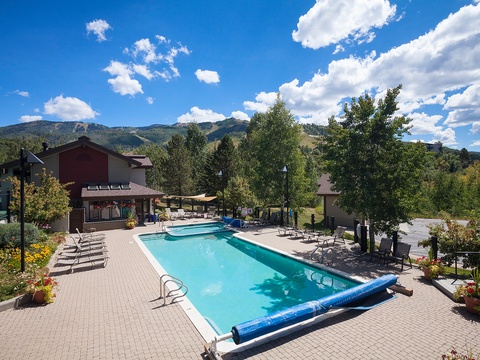 A scenic mountain resort features an inviting outdoor pool surrounded by lounge chairs, set against lush greenery and dramatic mountain peaks under a clear blue sky with scattered clouds.