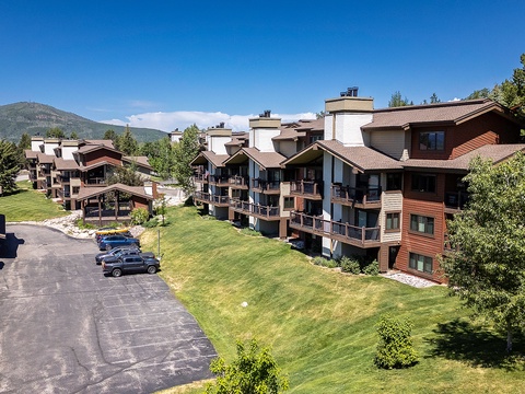 A mountain resort community featuring multi-level alpine-style condominiums with private balconies, expansive lawns, parking, and forested peaks beyond.