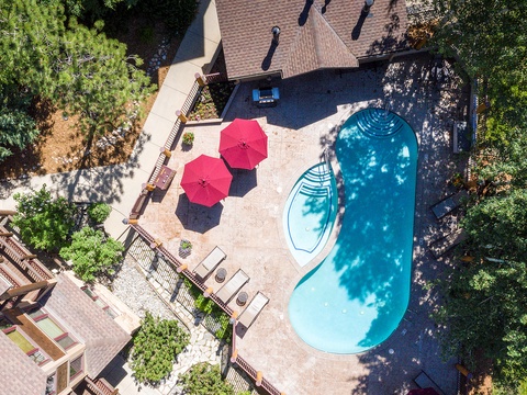 Aerial view of the property showcasing a kidney-shaped swimming pool, hot tub, patio with red umbrellas, and lush surrounding greenery.