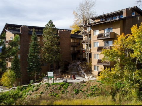 Multi-level property building surrounded by evergreens and golden aspens, nestled in a natural hillside setting with marked pathways leading to the entrance.