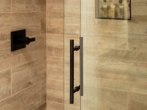 Glass shower door with modern black hardware awaits your refreshing morning routine in this sleek, travertine-tiled bathroom.