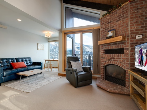 Your cozy living room features a warm brick fireplace, leather seating, and mountain views through the balcony door—perfect for relaxing after a day on the slopes.