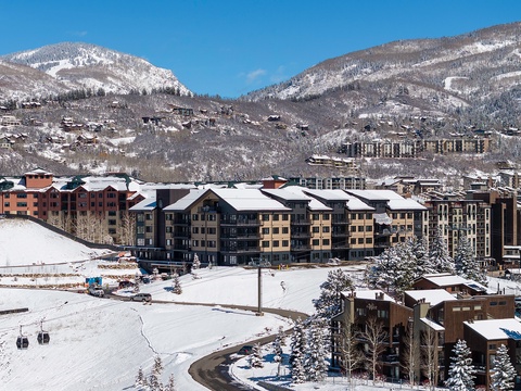 A snow-covered mountain resort village nestled in a valley, featuring multi-story lodges, ski lifts, and scenic alpine peaks under crisp blue skies.
