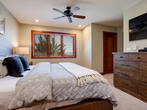 Unwind in your peaceful bedroom with plush bedding, warm wood accents, and serene garden views through the window. The ceiling fan and mounted TV ensure your comfort.
