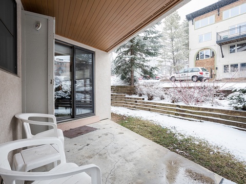 This covered terrace overlooks a snowy alpine landscape. White outdoor furniture provides a comfortable vantage point to enjoy the quiet winter setting, with evergreens and neighboring buildings adding to the seasonal charm.