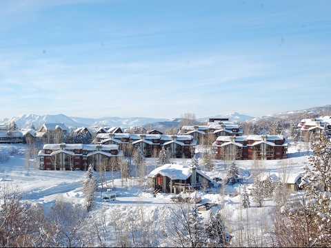 Snow-dusted vacation homes rest peacefully in a picturesque mountain valley, surrounded by pristine winter wilderness and distant peaks under clear skies.