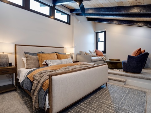 Your modern retreat features a plush bed layered with rich textures, warm wood beams overhead, and a cozy seating nook—all bathed in natural light for restful mornings and peaceful evenings.