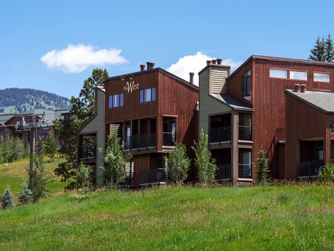 Alpine vacation property exterior featuring multi-level wood and siding architecture nestled against green ski slopes and mountain scenery under clear blue skies.