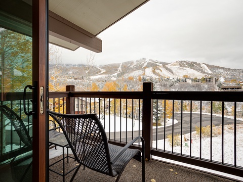 Snowy ski slopes overlook a balcony equipped with two chairs facing a mountain resort, framed by autumn-colored trees and covered terrain during an early winter transition.