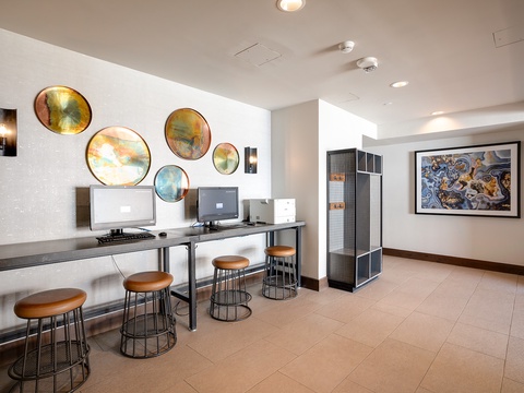 A modern business center featuring workstations with sleek desks, comfortable seating, and dual monitors. Vibrant circular art adorns the walls, creating an inspiring workspace for staying productive during your stay.