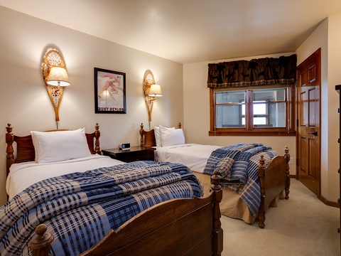 Cozy room with two plush beds draped in blue plaid throws, warm lighting from unique snowshoe sconces, and mountain views to greet you each morning.