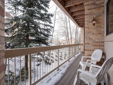 A snow-dusted private balcony with seating overlooks a quiet winter landscape of tall evergreens and neighboring buildings in this peaceful ski-area neighborhood.