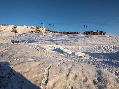 A scenic ski resort area with groomed slopes, ski lifts, and lodging facilities set against a clear blue winter sky, offering access to alpine winter sports.