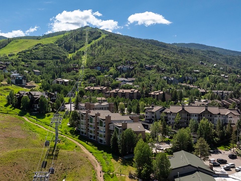 Aerial view of a mountain resort community featuring ski slopes, residential buildings, and forested hillsides during summer season with clear blue skies.